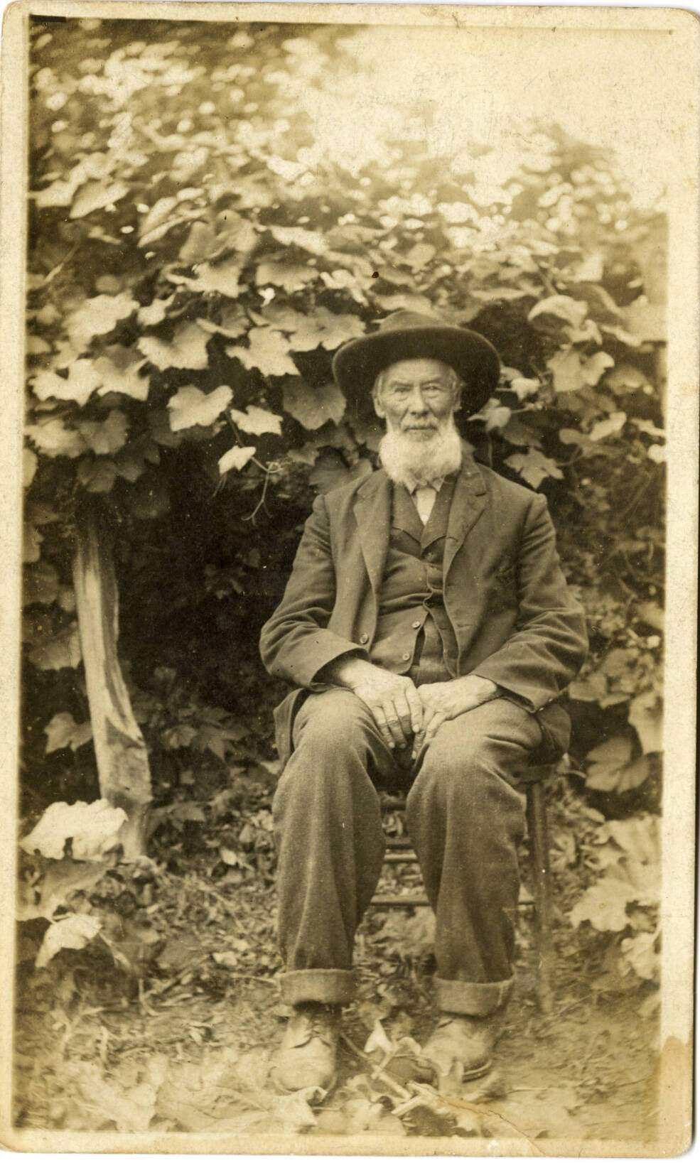 Historic photograph of a man sitting in a chair outside in front of a bush, wearing a dark suit and hat with a white beard.